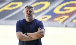 Mężczyzna z rękami skrzyżowanymi na piersi, w granatowej koszulce, stoi na tle stadionu. To trener Pogoni Szczecin.