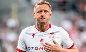 Piłkarz w białej koszulce z orłem, z ręką na sercu, wyraźnie przeżywa trudne chwile po kontuzji.