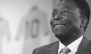 Uśmiechnięty Pele, legenda futbolu, w garniturze. W tle koszulka z numerem 10. Czy Pele żyje? Tak, jego dziedzictwo żyje wiecznie.