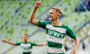 Radosny środkowy napastnik w zielono-białej koszulce świętuje bramkę na tle zielonych trybun stadionu.