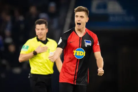 Piłkarz klubu piłkarskiego z Berlina, w czarno-czerwonej koszulce z logo Tedi, cieszy się po strzelonej bramce. Sędzia w żółtej koszulce stoi z tyłu.
