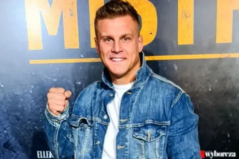 Uśmiechnięty Alan Kwieciński, którego wiek jest często przedmiotem dyskusji, w jeansowej kurtce.