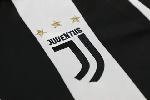 Zbliżenie na herb Juventusu z trzema złotymi gwiazdkami i napisem "JUVENTUS" na tle charakterystycznych czarno-białych pasów.