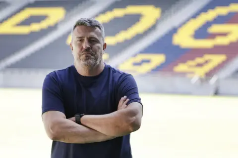 Mężczyzna z rękami skrzyżowanymi na piersi, w granatowej koszulce, stoi na tle stadionu. To trener Pogoni Szczecin.