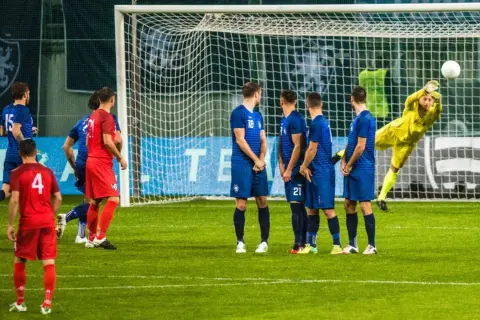 Piłkarze w niebieskich strojach czekają na wznowienie gry. Bramkarz w żółtym stroju broni strzał. Czas na przerwę w meczu?