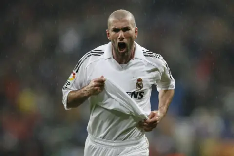 Zinedine Zidane w białej koszulce Realu Madryt, z podniesioną koszulką, krzyczy radośnie do kibiców.