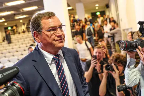 Mężczyzna w garniturze, otoczony przez fotoreporterów z aparatami, przypomina jednego z kapitanów reprezentacji Polski, gotowego na ważne wydarzenie.