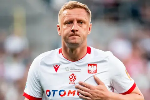 Piłkarz w białej koszulce z orłem, z ręką na sercu, wyraźnie przeżywa trudne chwile po kontuzji.