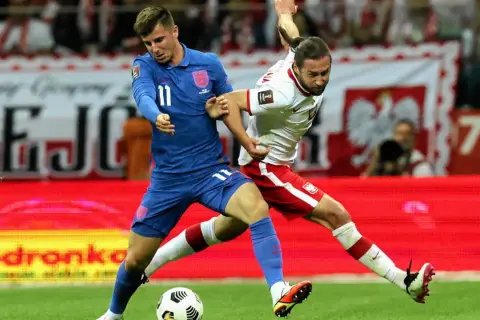 Piłkarz Anglii w niebieskiej koszulce walczy o piłkę z zawodnikiem Polski w białej. Emocjonujący mecz Anglia Polska.
