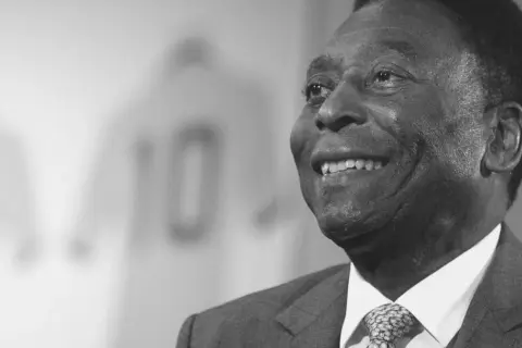 Uśmiechnięty Pele, legenda futbolu, w garniturze. W tle koszulka z numerem 10. Czy Pele żyje? Tak, jego dziedzictwo żyje wiecznie.