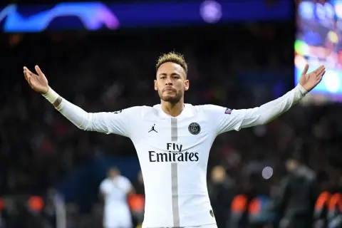 Neymar w białej koszulce PSG, z charakterystycznymi fryzurami, unosi ręce na tle tłumu.