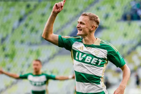 Radosny środkowy napastnik w zielono-białej koszulce świętuje bramkę na tle zielonych trybun stadionu.