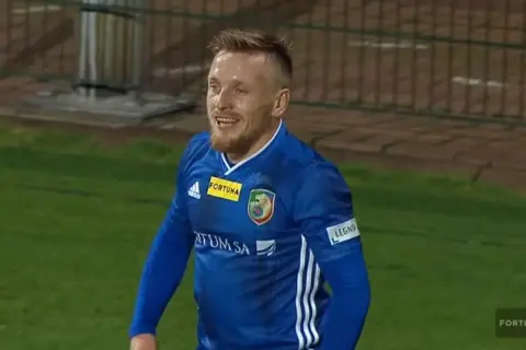 Uśmiechnięty piłkarz w niebieskiej koszulce, być może najniższy piłkarz na świecie, cieszy się grą na boisku.