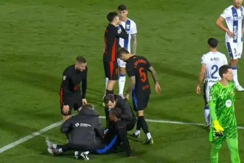 Piłkarz leży na boisku, udzielana jest mu pomoc medyczna. Widać obawy o jego stan po kontuzji.