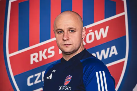 Łysy mężczyzna, trener Rakowa, patrzy prosto przed siebie na tle klubowego herbu.
