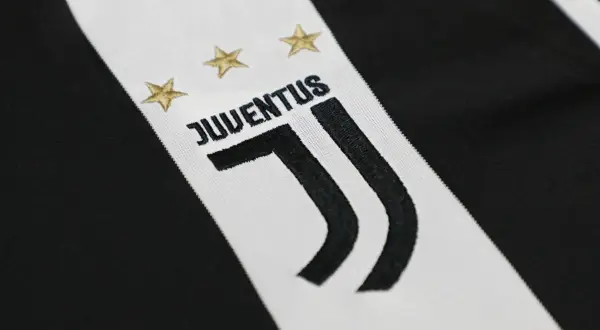 Zbliżenie na herb Juventusu z trzema złotymi gwiazdkami i napisem "JUVENTUS" na tle charakterystycznych czarno-białych pasów.