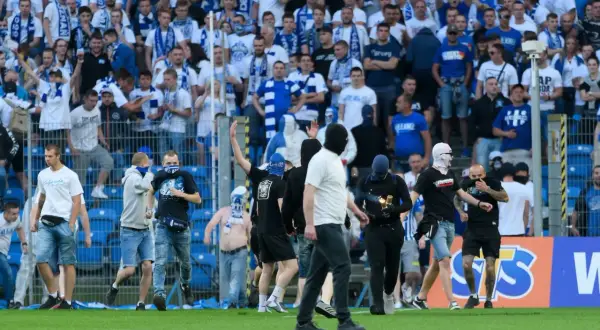 Kibice Lecha Poznań w niebiesko-białych barwach, niektórzy w kominiarkach, na murawie stadionu.