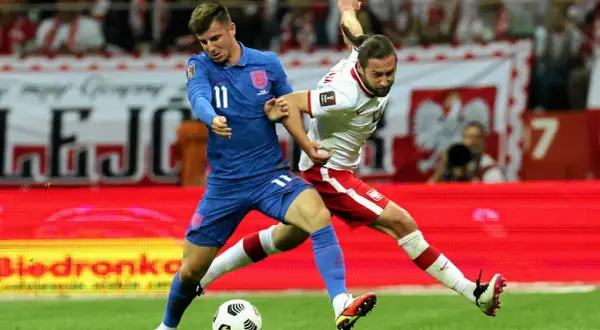 Piłkarz Anglii w niebieskiej koszulce walczy o piłkę z zawodnikiem Polski w białej. Emocjonujący mecz Anglia Polska.