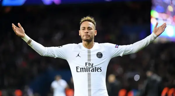 Neymar w białej koszulce PSG, z charakterystycznymi fryzurami, unosi ręce na tle tłumu.
