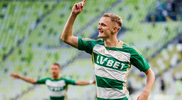 Radosny środkowy napastnik w zielono-białej koszulce świętuje bramkę na tle zielonych trybun stadionu.
