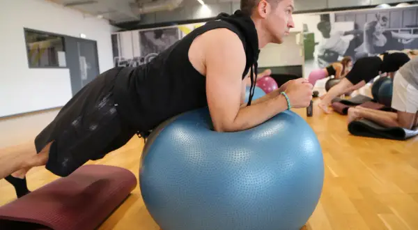 Mężczyzna w czarnej bluzie i spodenkach ćwiczy na dużej, niebieskiej piłce fitness.