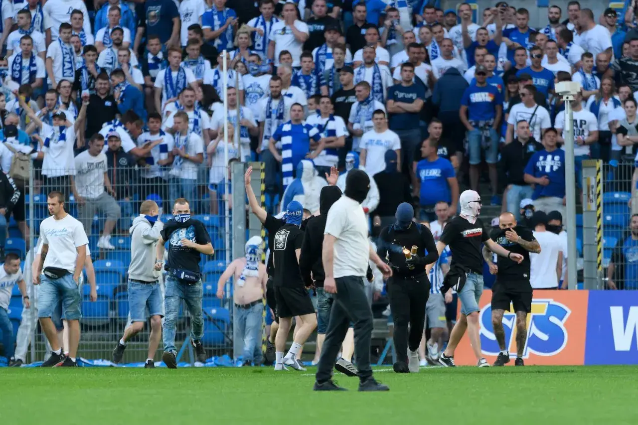 Kibice Lecha Poznań w niebiesko-białych barwach, niektórzy w kominiarkach, na murawie stadionu.