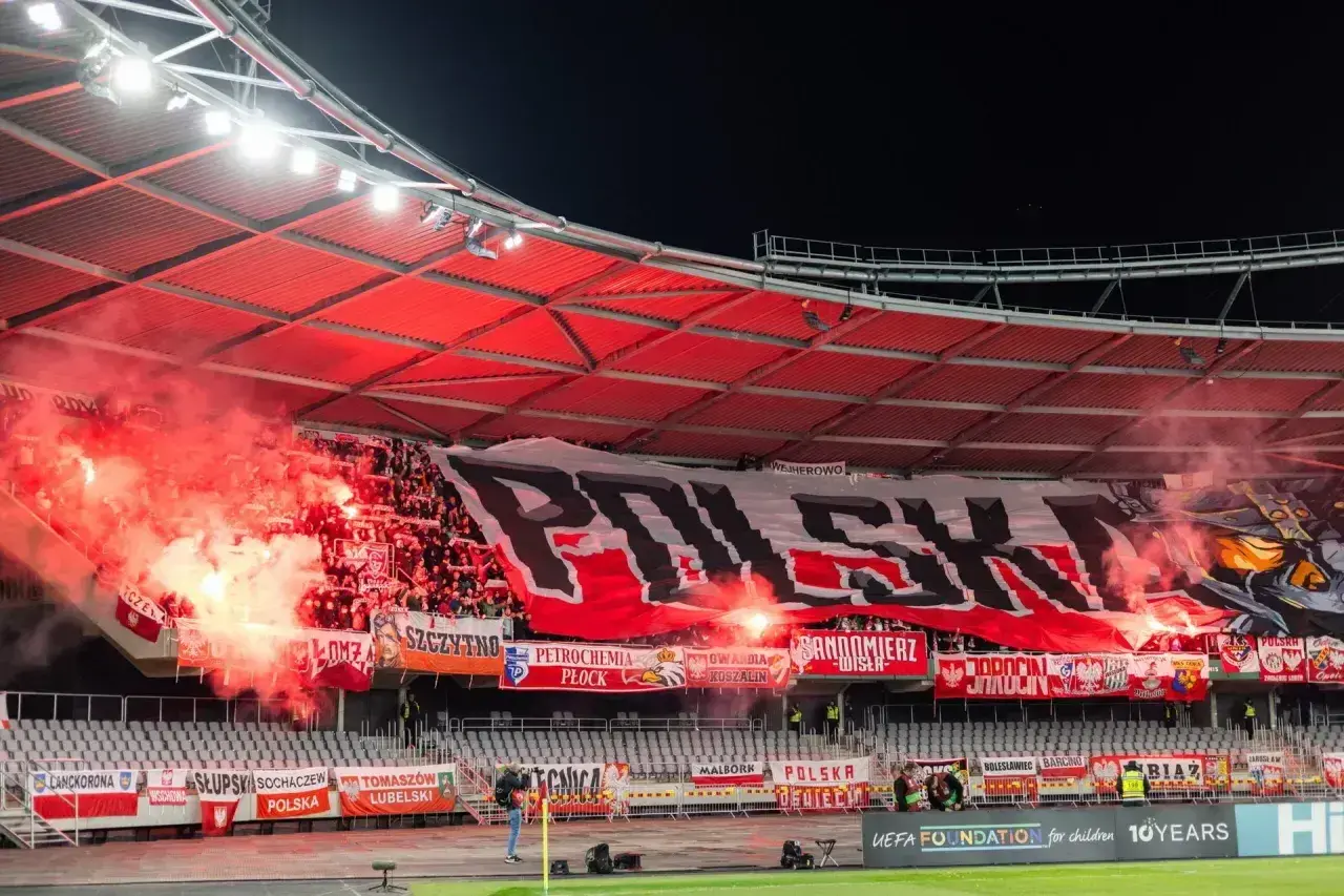 Kibice na stadionie odpalają race, tworząc czerwony dym. Na trybunach rozwinięty ogromny transparent "POLSKA" i flagi.