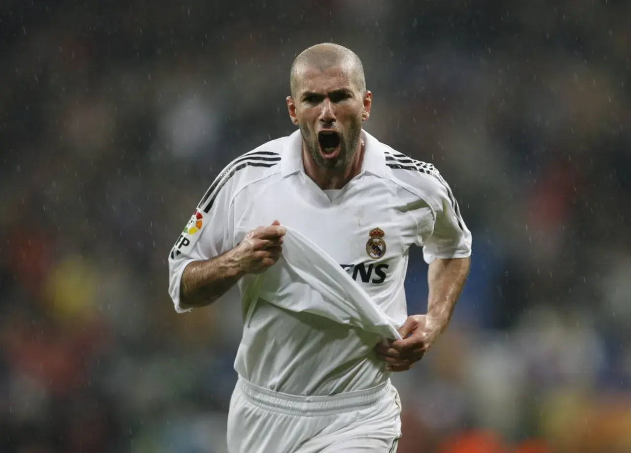 Zinedine Zidane w białej koszulce Realu Madryt, z podniesioną koszulką, krzyczy radośnie do kibiców.