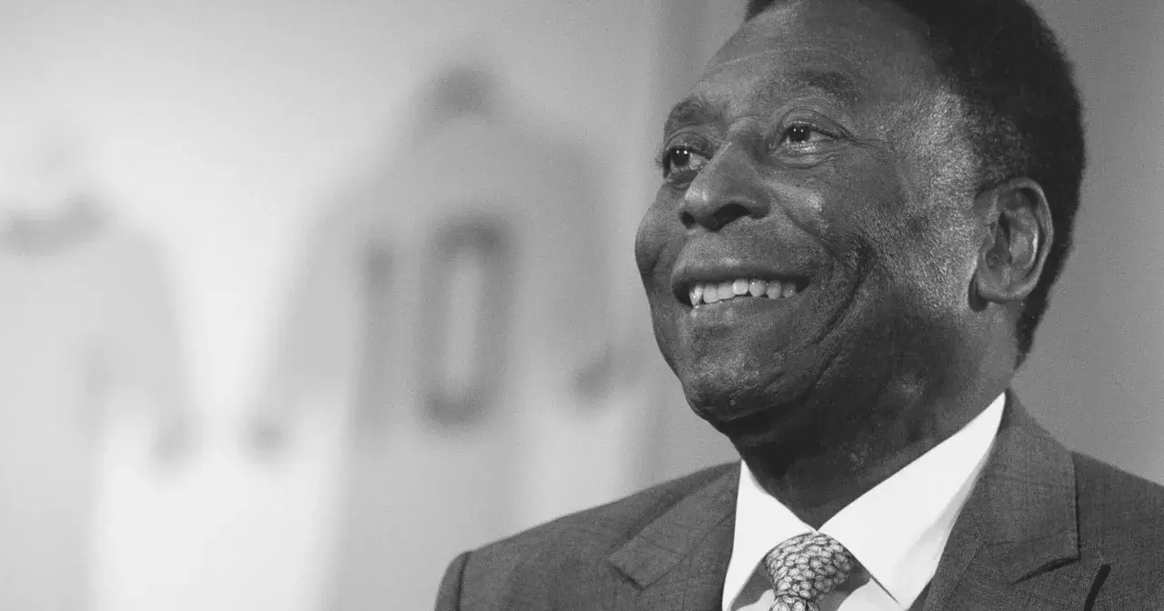 Uśmiechnięty Pele, legenda futbolu, w garniturze. W tle koszulka z numerem 10. Czy Pele żyje? Tak, jego dziedzictwo żyje wiecznie.