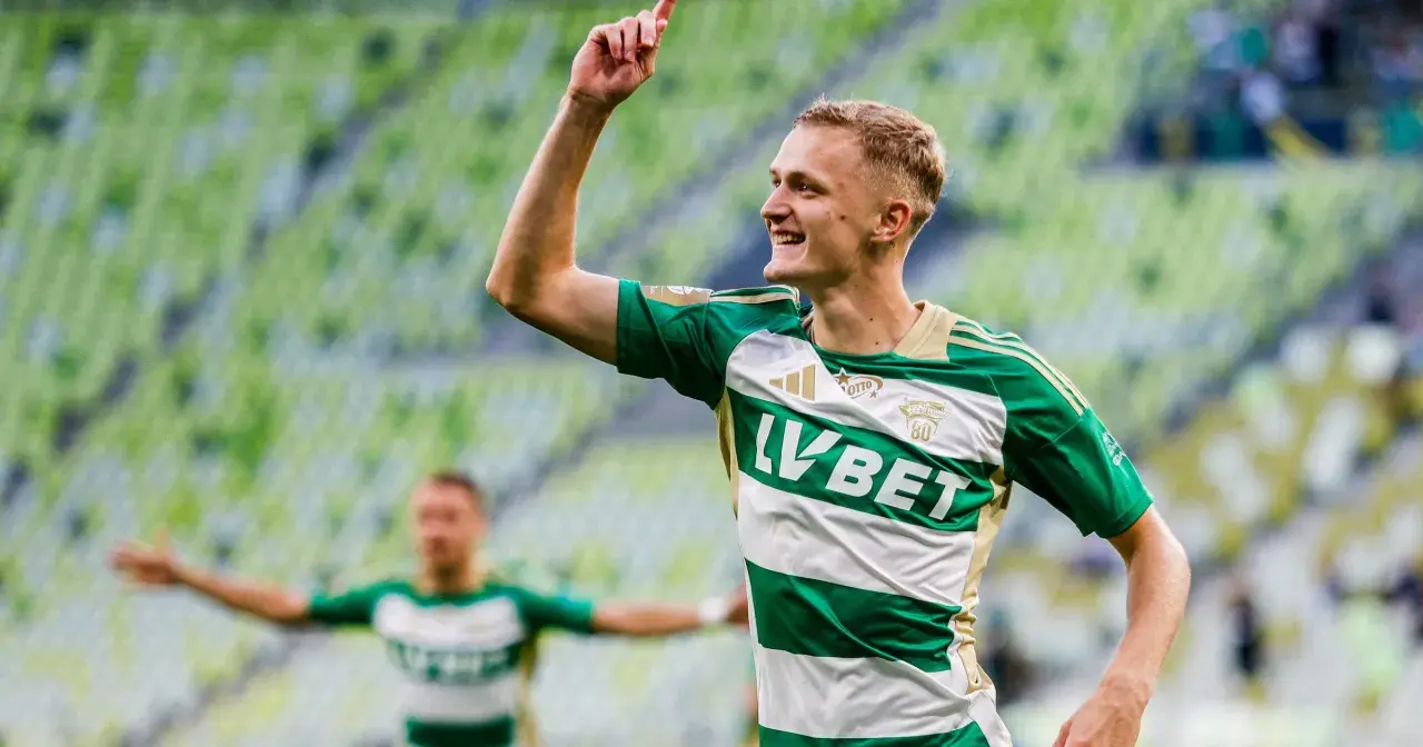 Radosny środkowy napastnik w zielono-białej koszulce świętuje bramkę na tle zielonych trybun stadionu.
