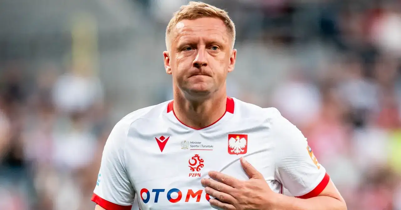 Piłkarz w białej koszulce z orłem, z ręką na sercu, wyraźnie przeżywa trudne chwile po kontuzji.