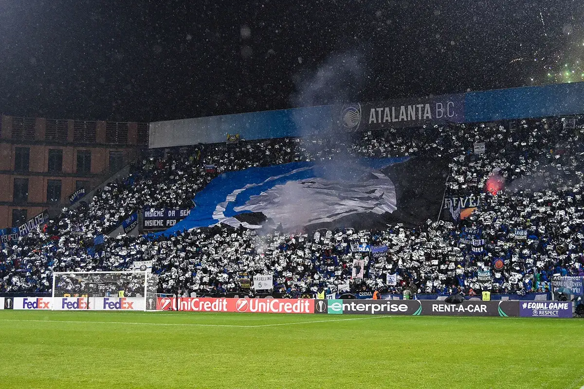 Trybuny stadionu wypełnione kibicami włoskiego klubu piłkarskiego z Bergamo. Fani tworzą oprawę z niebiesko-czarnym transparentem i racami.
