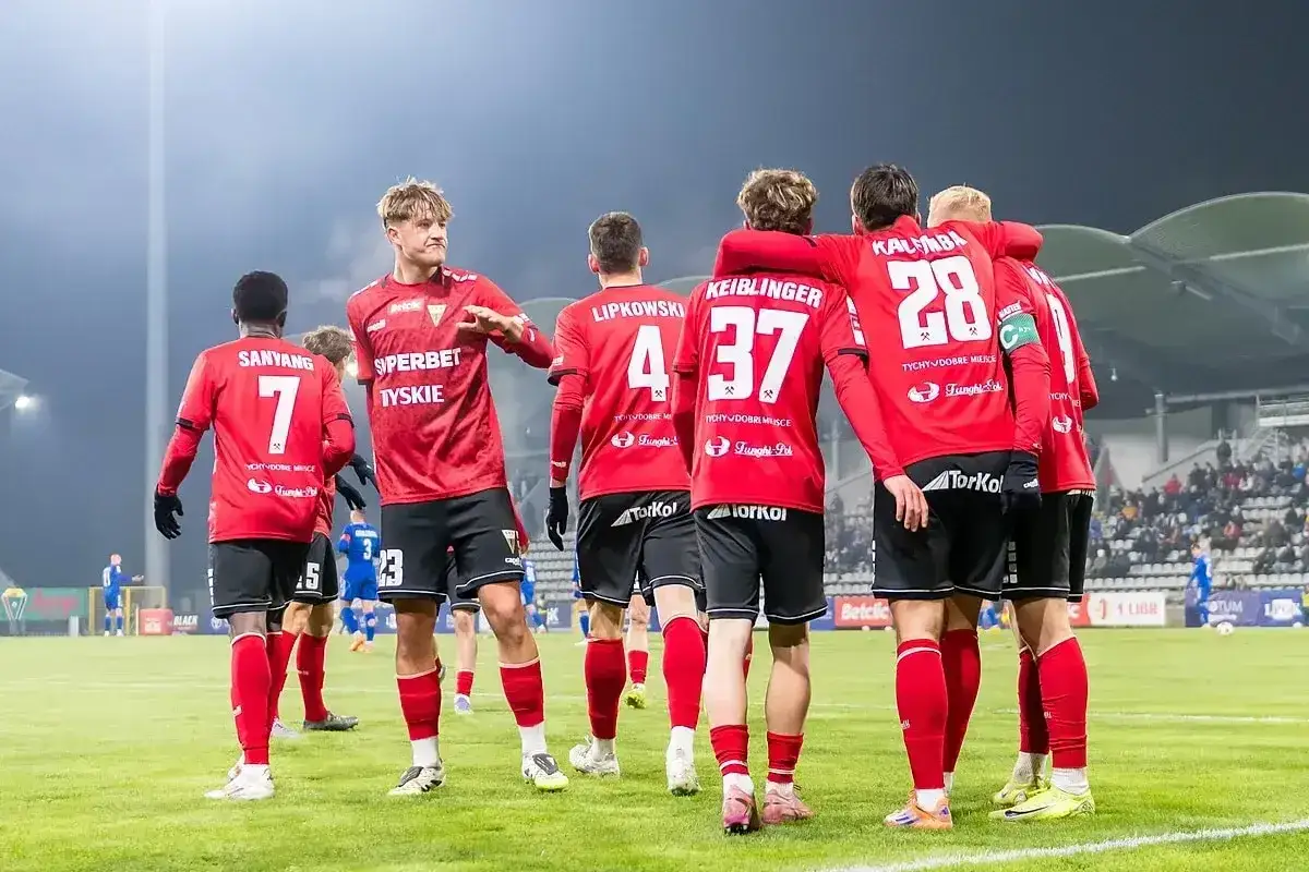 Piłkarze GKS Tychy w czerwonych strojach świętują na boisku. Kibice i stadion w tle.