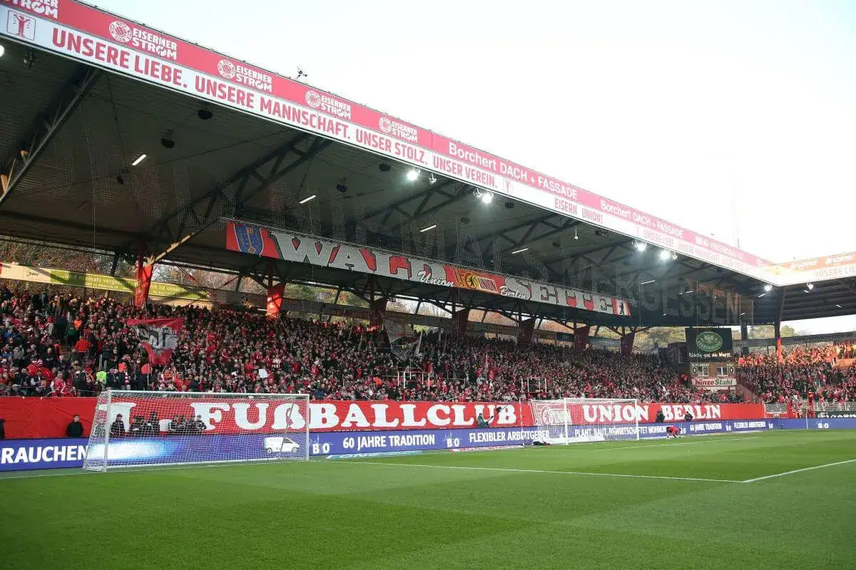 Trybuna stadionu wypełniona kibicami klubu piłkarskiego z Berlina. Na banerach widnieje napis