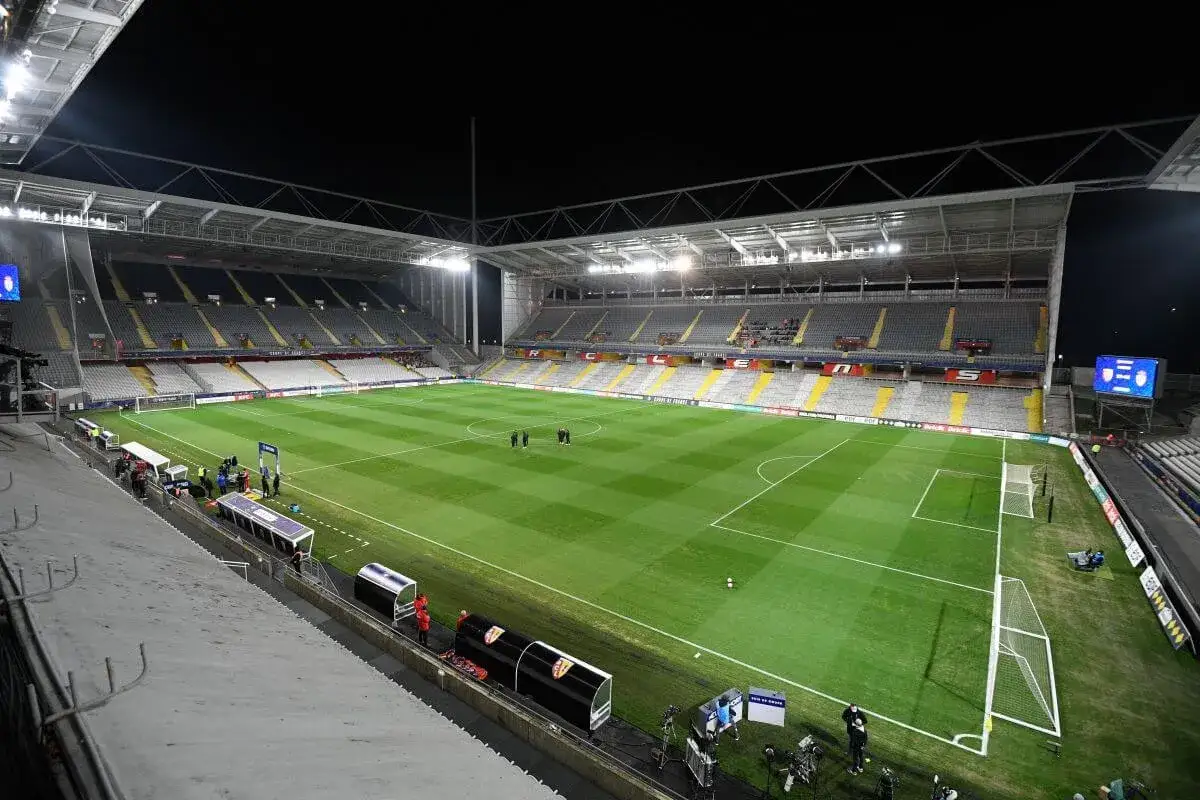 Piłkarski stadion w nocy, z pustymi trybunami i oświetloną murawą. Widać ławki rezerwowych i bandy reklamowe z napisami sugerującymi, że to obiekt w Belgii lub Francji.