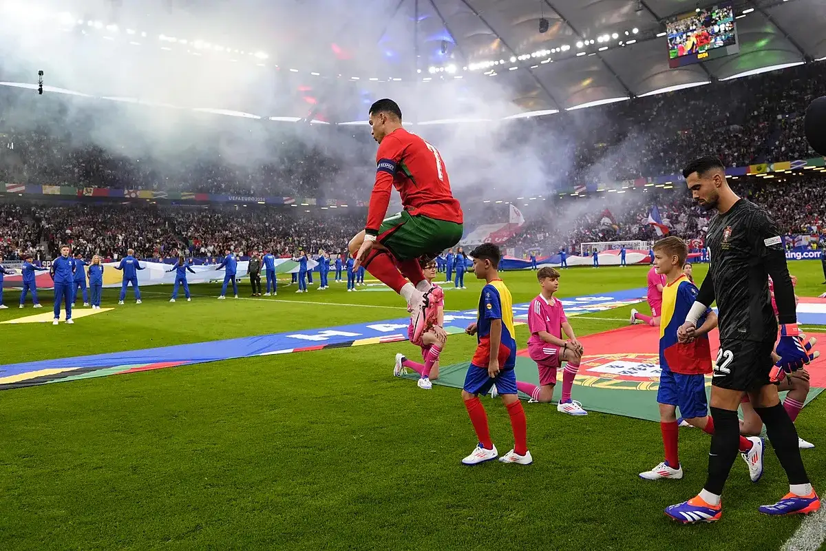 Piłkarz w skoku na tle stadionu, gdzie trwa mecz Portugalia Chorwacja. Dzieci w strojach sportowych i bramkarz w tle.